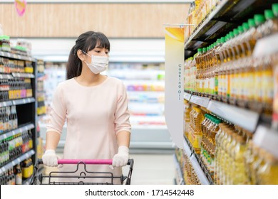 Asian Woman With Hygienic Mask And Rubber Glove With Shopping Cart In Grocery And Looking For Cooking Oil Bottle To Buy During Covid-19 Outbreak For Preparation For A Pandemic Quarantine