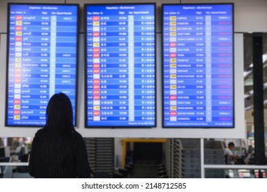 An Asian Woman In An International Airport Looks At A Flight Information Board, Holding A Passport In Her Hand, Checking Her Flight. Travel Holiday