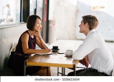 An Asian Woman Is Interviewing A Young Caucasian White Man For An Internship Job In Asia. They Are Sitting At A Desk In The Shade Outside In The Day And Talking Over Coffee. 