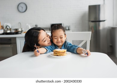 Asian woman kissing smiling daughter near pancakes at home - Powered by Shutterstock