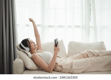 Asian woman is laying on a couch with her headphones on and a cell phone in her hand. She is enjoying her music and is in a relaxed state - Powered by Shutterstock