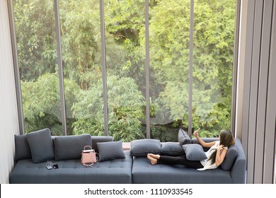 Asian Woman Laying Sitting And Taking Selfie On Sofa Near Big Glass Windows, Relaxing Alone In House Or Apartment With Green Forest In Background, Concept For People Who Living In Modern Lifestyle.