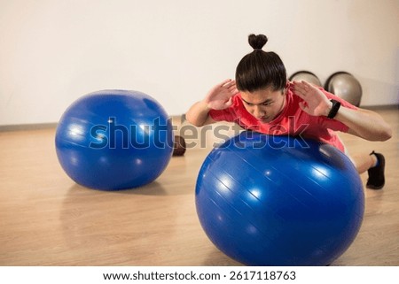 Asian woman leaning forward on blue stability ball in fitness studio, building core strength. Exercise, balance, stability, workout, gym, minimalist, wellness