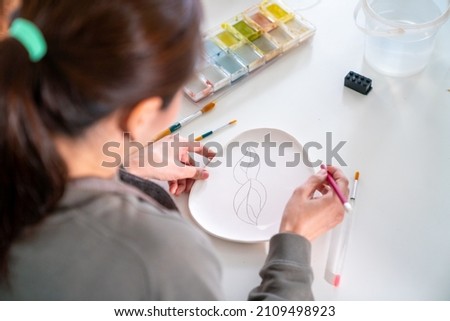 Asian woman learning color painting her self-made pottery plate at home. Confidence female relax and enjoy handicraft activity lifestyle hobbies ceramic sculpture painting workshop at pottery studio