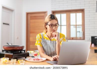 Asian woman learns to cook, she watches video recipes on a laptop in the kitchen and cook a dish from the beef steak. learning online cooking at home concept - Shutterstock ID 2030573891