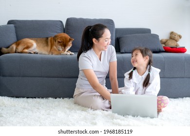 Asiatische Frauen und kleine Mädchen genießen gemeinsam im Wohnzimmer mit Laptop und sitzen auf dem Boden, während ihr Shiba Hund auf dem Sofa liegt.