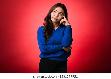 An Asian woman with long brown hair, wearing a blue sweater, poses with a thoughtful expression, touching her temple with her finger. She stands against a red background - Powered by Shutterstock