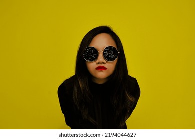 Asian Woman With Long Brunette Hair, Dark Round Shades With TV Static, Red Lips, And Black Blouse Leans Forward Against Bold Yellow Background. Symbol For Social Media, Fame, And Celebrity Obsession.