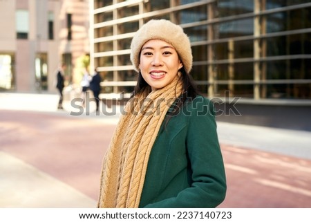 An Asian woman looking at the camera outside an office area with business people in the background. Cheerful Chinese girl in winter clothes in the city. High quality photo