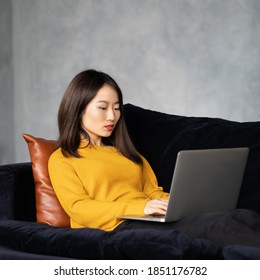 Asian Woman Looking At Laptop, Working At Home, Liying On Sofa. Unhappy, Upset Japanese Female Freelancer Smiling And Typing At Computer In Hotel On Couch