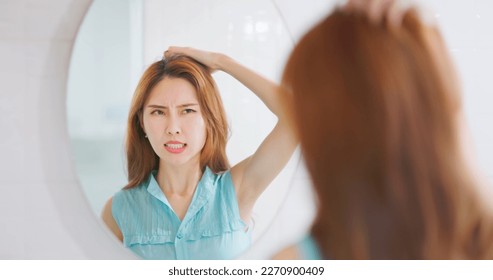 Asian woman looking in mirror is scratching head from itching and having messy oily hair - Shutterstock ID 2270900409