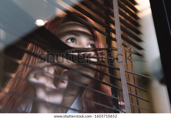 Asian woman looking through window blinds spying
on neighbours - Young lonely millennial woman peeping through glass
observing gossip and action outdoors - introvert, privacy and
intrusive concepts
