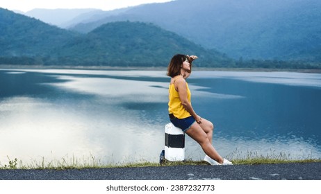 Asian Woman looking at view sitting near on the lake shore at dam. Chilling woman relaxing on sunny lakeside dock. Against blurred background of green nature mountain.