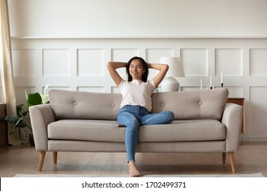 Asian woman looks at distance resting leaned on couch enjoy fresh air in summer day in modern fashionable living room interior, full length image. Contemporary apartments owner or carefree day concept