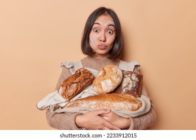 Asian woman looks surprisingly at camera holds napkin with fresh bread works in bakehouse keeps lips rounded wears casual jumper apron isolated over beige background. Bakery and food concept - Powered by Shutterstock