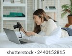 Asian woman lying on bed, happy working at home and holding camera, checking photos, contributors selling, stock photos on website, working online in apartment