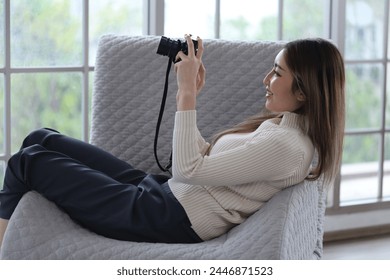 Asian woman lying on sofa holding DSLR camera, reviewing photos from camera before sending photos for sale in apartment - Powered by Shutterstock