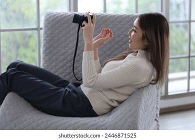 Asian woman lying on sofa holding DSLR camera, reviewing photos from camera before sending photos for sale in apartment - Powered by Shutterstock
