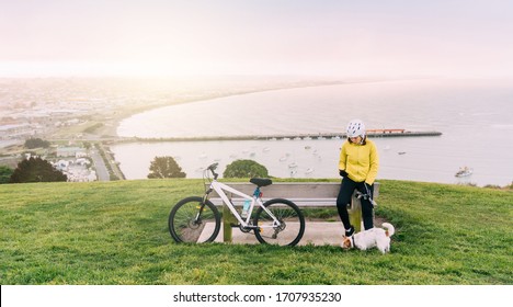 Asian Woman Making Uphill With Mountain Bike. A Woman Using A Cell Phone At Lookout Point With A Dog At Oamaru, New Zealand.