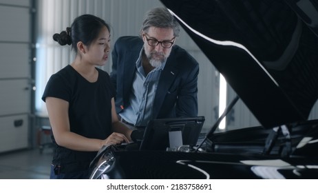 An Asian Woman Mechanic Consulting A Man Client In A Car Service, Swiping On Tablet And Diagnosing A Car With A Breakdown