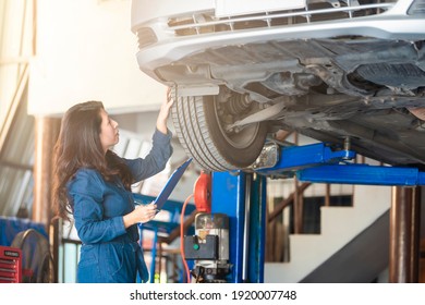 Asian Woman Mechanic Holding A Clipboard And Repairs The Brakes Of An Automobile With Tire On A Hydraulic Lift.