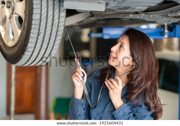 Asian woman mechanic is listen to\
drive shaft of old car, wheel and joint of steering rack for check\
service by ears phones doctor for service in\
garage.