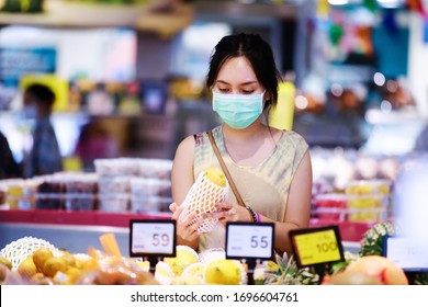 Asian Woman In Medical Face Mask Chooses Fruits While Shopping In Supermarket. Covid-19 Spreading Outbreak 