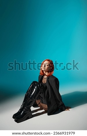 asian woman with nose piercing sitting in oversized blazer and black latex boots on blue backdrop
