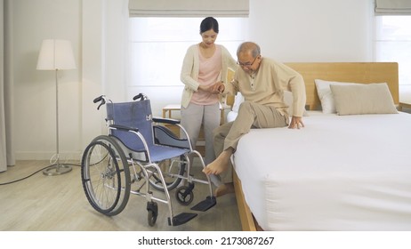Asian Woman Nurse, Daughter Help Father To Get Up From Bed To Wheelchair, Supporting Old Senior Elderly Patient In Bedroom In Home Or House In Healthcare. People Lifestyle. Family Disability Therapy.