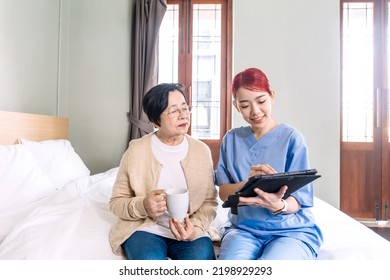 Asian Woman Nurse Wearing Scrubs Report The Health Status Of Senior Asian Woman With Digital Tablet In The Bedroom. Caregiver Visit At Home. Home Health Care And Nursing Home Concept.