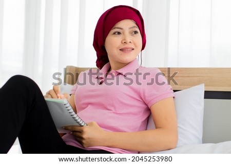 Asian woman on pink shirt, black trousers, cover head by red scarf as breast cancer patient, up knee to take sad note on paper while relaxing on house bed beside alarm clock, photo frame, plant pot