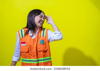 An Asian woman in an orange safety vest smiles brightly with her eyes closed, playfully shielding her face with her hand. She stands against a vibrant yellow background, exuding joy and positivity. - Powered by Shutterstock