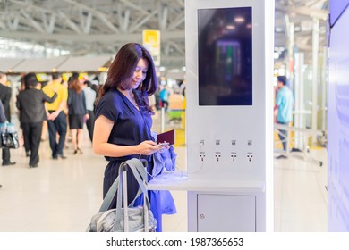 Asian woman with passport, suitcase standing at a charging station and looking at her smartphone. Recharging mobile phones from free charge station at airport.