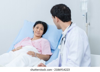 Asian Woman Patient Lying Down On Bed With Doctor In Hospital Ward. Physician Therapist Pharmacist Man Visit And Give Diagnosis Service Treatment Sick Female With Happiness And Smiling At Hospital.