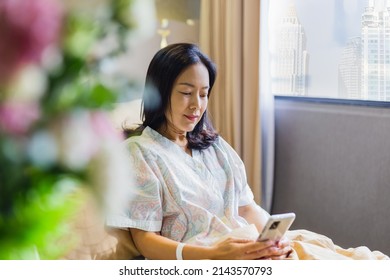 Asian Woman Patient Smiling While Sit On Bed Making Video Call On Cell Phone In Hospital Ward