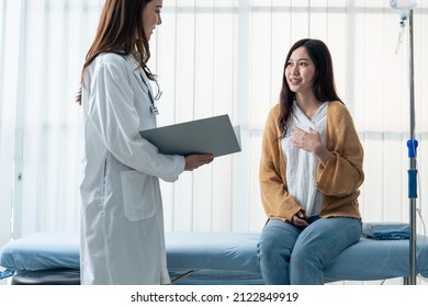 Asian Woman Patient Visit And Consult Health Problem With Young Doctor. Attractive Beautiful Female Therapist Practitioner Working In Office Hospital To Give Advice Treatment To Sick Girl Sit On Bed.