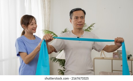 Asian Woman Personal Care Attendant Assisting Older Male Working Out With Elastic Band At Home. Social Homecare And Senior Fitness Concept