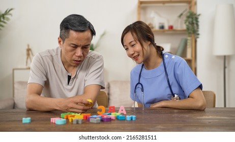 Asian Woman Physiotherapist Guiding Older Alzheimer’s Patient Through Rehabilitation Training At Home. He Looks At Letter Blocks Trying To Recognize Them