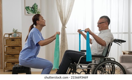 Asian Woman Physiotherapist Teaching Handicapped Older Man How To Use Elastic Band While She Is Doing Resistance Training On Wheelchair At Home