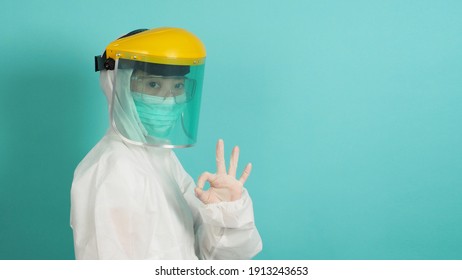 Asian Woman In PPE Suit , Face Shiled,medical Gloves And Wear Face Mask  Do OK Hand Sign On Green, Tiffany Blue Background.