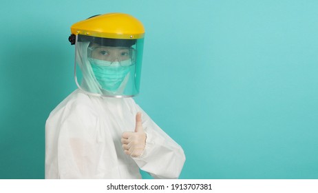 Asian Woman In PPE Suit , Face Shiled,medical Gloves And Wear Face Mask  Do Thumb Up Or Like Hand Sign On Green, Tiffany Blue Background.