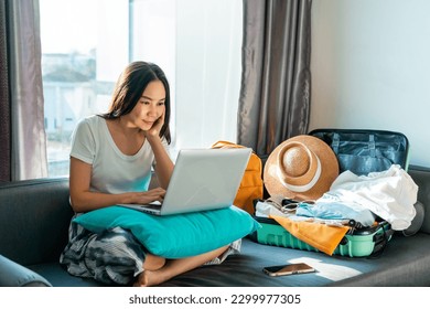 Asian woman prepare things into baggage for vacation trip at home. Happy female using computer to planning travel trip and booking tickets or hotel room on web. Travel, summer and holiday concept.
