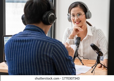 Asian woman radio hosts gesturing to microphone while interviewing a man guest in a studio while recording podcast for online show in studio together.