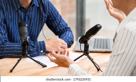 Asian Woman Radio Hosts Gesturing To Microphone While Interviewing A Man Guest In A Studio While Recording Podcast For Online Show In Studio Together.