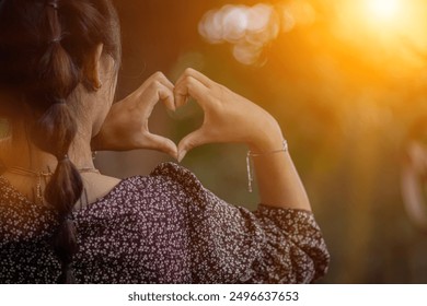 An Asian woman raises her hand to show a heart-shaped symbol signifying love, friendship and sympathy on a blurry background of a green forest looking beautiful. friendship and love concept - Powered by Shutterstock