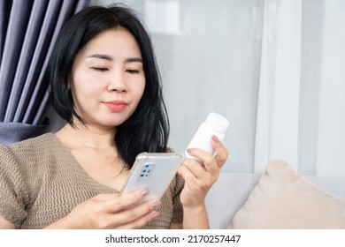 Asian Woman Reading A Prescription Medication Label Hand Holding Bottle Of Drug And Checking Information On Mobile Phone  