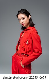 asian woman in red jacket decorated with brooch and glove standing with hands in pockets isolated on grey - Shutterstock ID 2235546679