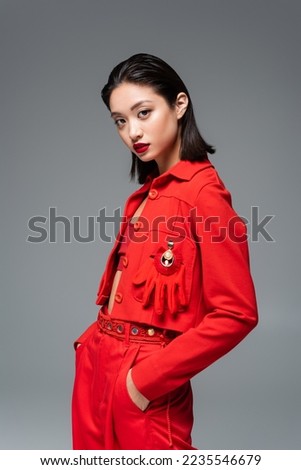 asian woman in red jacket decorated with brooch and glove standing with hands in pockets isolated on grey