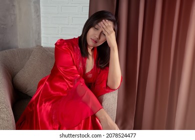 Asian woman in a red satin nightgown and red long full length robe with headache, she is sitting on the gray sofa and touching her forehead. Thai woman dizzy at night.
