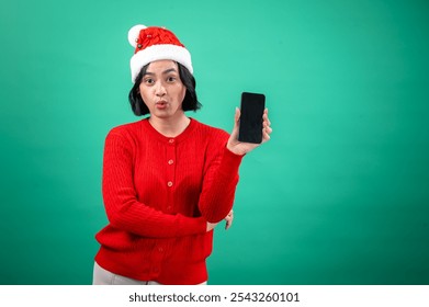 An Asian woman in a red sweater and Santa hat stands against a green background, smiling brightly as she holds up a smartphone in her right hand, showcasing its blank screen. - Powered by Shutterstock